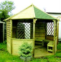 Drewniana altana ogrodowa z sześciokątnym dachem, częściowo pokrytym zieloną dachówką, z dekoracyjnymi krzyżowymi kratami i wbudowaną ławką, otoczona zielenią.